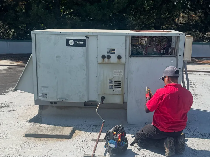 A Dayco technician in a red shirt kneeling to service a Trane rooftop unit