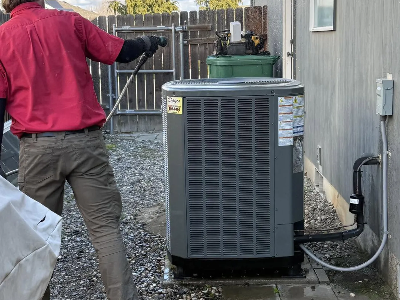 A Dayco technician in a red company shirt standing beside an outdoor air conditioner
