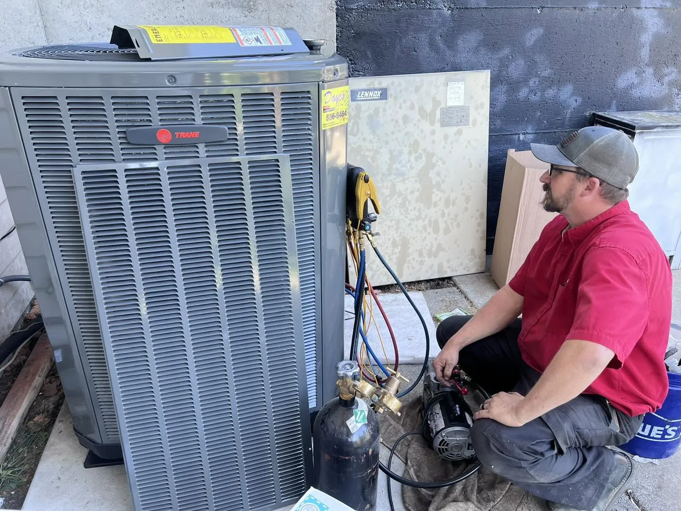 A Dayco technician in a red shirt kneeling beside a new Trane outdoor unit with gauges, a vacuum pump and a nitrogen tank connected