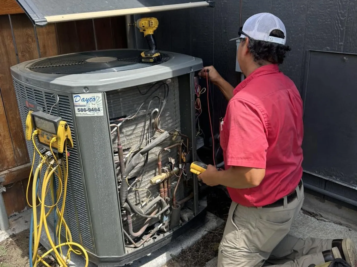 A Dayco technician in a red shirt servicing an outdoor Trane unit beside a house
