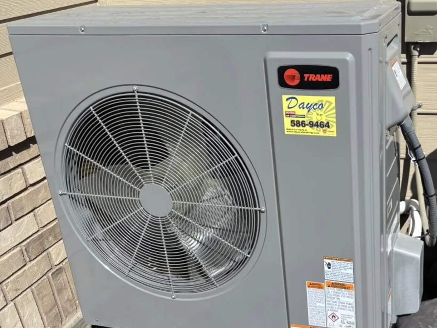 A new Trane outdoor unit with a Dayco service sticker, installed against a brick house