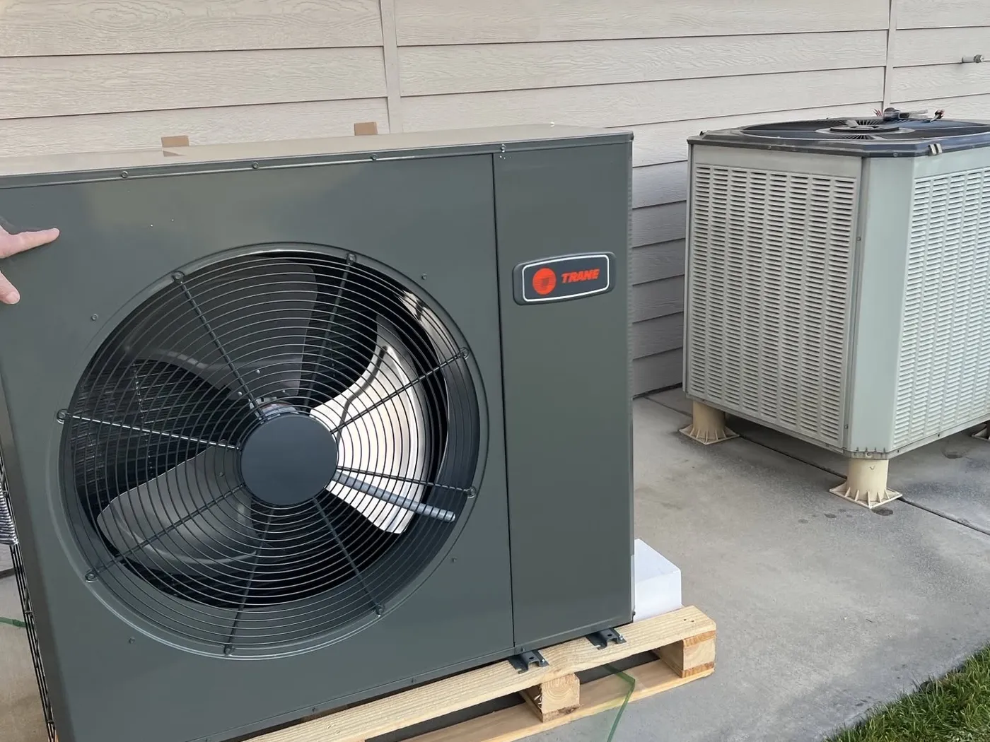 A new Trane heat pump on its shipping pallet beside a house, next to the older outdoor unit it is replacing